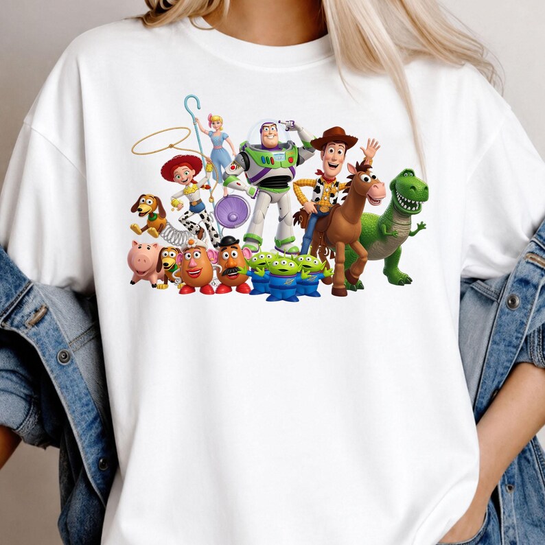 Może przedstawiać: Biały t-shirt z kr&oacute;tkim rękawem z kolorowym nadrukiem graficznym przedstawiającym r&oacute;żne postacie z Toy Story, w tym Chudego, Buzza Astrala, Jessie, Rexa, Cienkiego, Hamm, Pana i Panią Bulwę, Pastereczkę, Celka oraz zielonych kosmit&oacute;w.