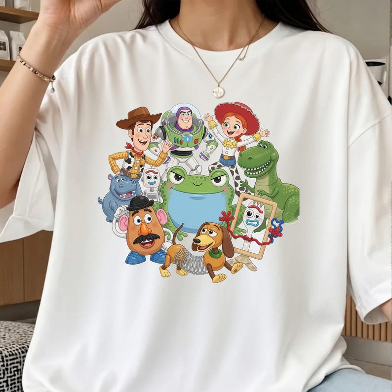 Może przedstawiać: Biały t-shirt z kr&oacute;tkim rękawem z kolorowym nadrukiem graficznym przedstawiającym r&oacute;żne postacie animowane, w tym kowboja, strażnika kosmosu, kowbojkę, zielonego dinozaura, postać ziemniaka oraz psa ze sprężyną zamiast tułowia.