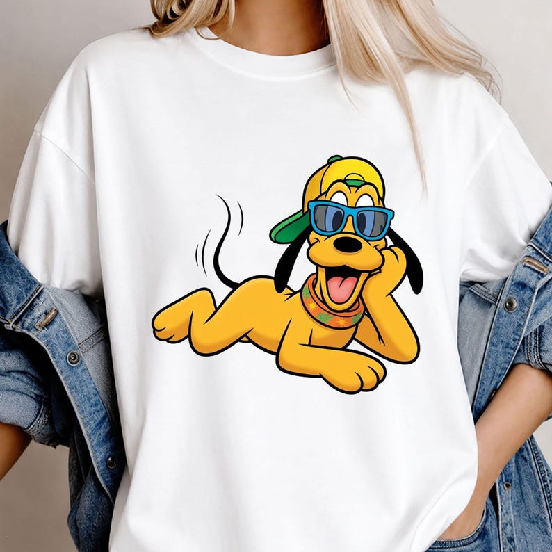May include: A white t-shirt featuring a graphic of a yellow cartoon dog wearing a green and yellow baseball cap, blue sunglasses, and a colorful patterned collar. The dog is lying down with its head resting on one paw, smiling with its tongue out.