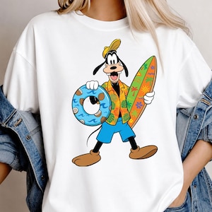 May include: A white t-shirt featuring a graphic of the cartoon character Goofy wearing a yellow cap, an orange floral shirt, and blue shorts. Goofy is holding a blue coconut-patterned swim ring in one hand and a colorful surfboard in the other.