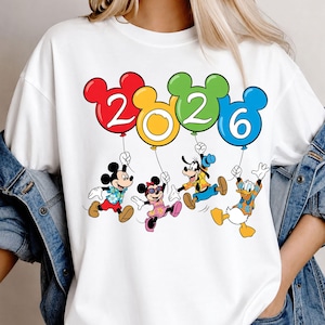 May include: A white t-shirt featuring a graphic of Mickey Mouse, Minnie Mouse, Goofy, and Donald Duck holding four colorful balloons shaped like Mickey Mouse heads. The balloons display the year 2026 in white text against red, yellow, green, and blue backgrounds.