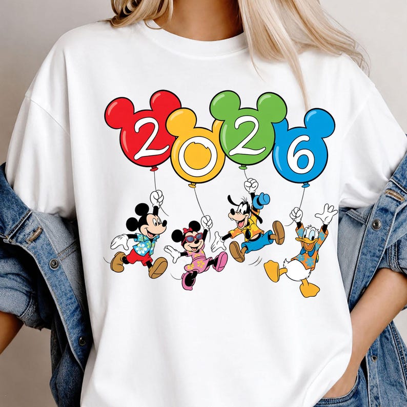 May include: A white t-shirt featuring a graphic of Mickey Mouse, Minnie Mouse, Goofy, and Donald Duck holding four colorful balloons shaped like Mickey Mouse heads. The balloons display the year 2026 in white text against red, yellow, green, and blue backgrounds.