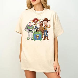 Może przedstawiać: Beżowy t-shirt z kr&oacute;tkim rękawem z nadrukiem graficznym przedstawiającym Buzza Astrala, Jessie, Celka i Chudego z Toy Story. Pod postaciami znajduje się zielony znak w kształcie żaby z napisem Toy Story 5 wydrukowanym ż&oacute;łtymi i czerwonymi literami.
