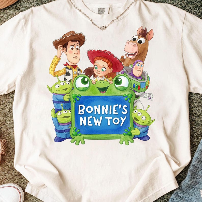 Może przedstawiać: Kremowy t-shirt z nadrukiem graficznym przedstawiającym postacie z Toy Story, w tym Chudego, Jessie, Bystrego, Buzza Astrala i trzy zielone ufoludki otaczające zielony znak w kształcie żaby z białym napisem BONNIE'S NEW TOY.