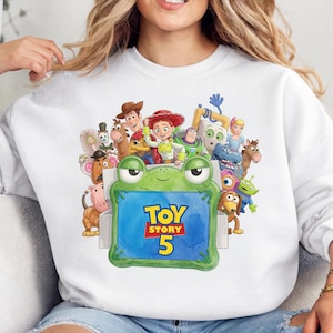 Op de afbeelding: Een wit sweatshirt met ronde hals en een kleurrijke grafische print van diverse Toy Story-personages rondom een groen, kikker-vormig kader. In het midden van het kader staat de blauwe tekst Toy Story 5 in een vetgedrukt geel en rood lettertype.