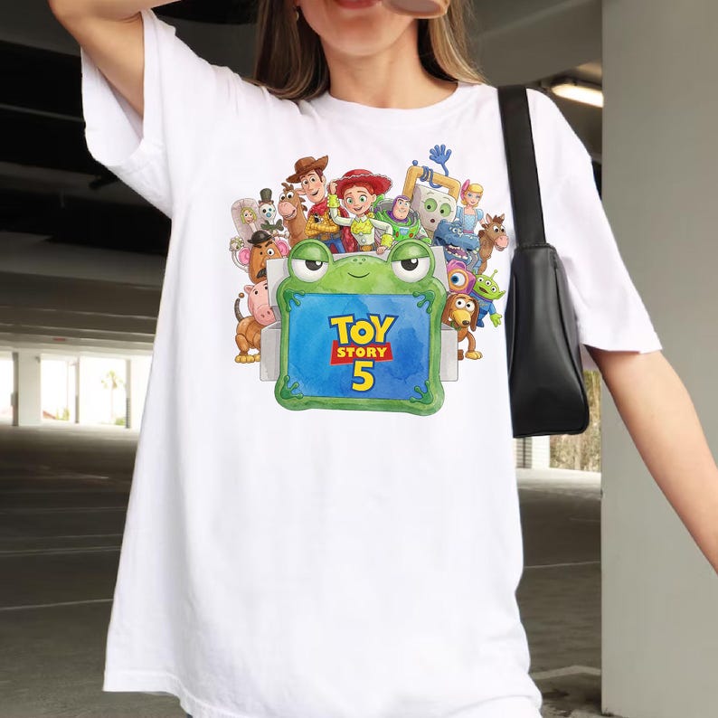 Może przedstawiać: Biały t-shirt z kr&oacute;tkim rękawem z kolorowym nadrukiem graficznym przedstawiającym r&oacute;żne postacie z Toy Story otaczające zieloną żabę trzymającą niebieski znak z napisem TOY STORY 5 w ż&oacute;łtym i czerwonym kolorze. Okrągły dekolt i luźny kr&oacute;j.