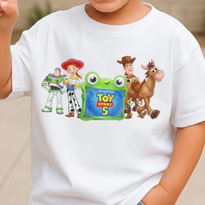 Może przedstawiać: Biały t-shirt z nadrukiem graficznym przedstawiającym Buzza Astrala, Jessie, Chudego i Bystrego z Toy Story. Pośrodku znajduje się zielona ramka w kształcie żaby z napisem Disney PTIAA TOY STORY 5 niebieskimi i ż&oacute;łtymi literami.
