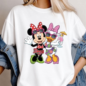 May include: A white short-sleeved t-shirt featuring a graphic print of Minnie Mouse and Daisy Duck wearing sunglasses and tropical-themed swimwear. Minnie wears a red polka-dot top and floral skirt, while Daisy holds a pink cocktail with an umbrella.