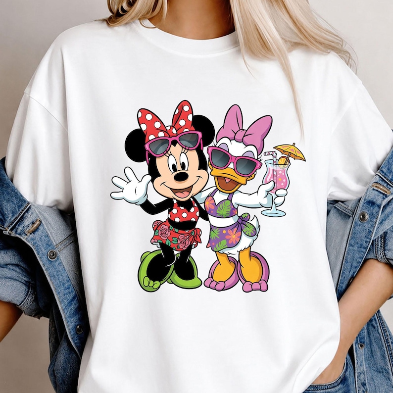 May include: A white short-sleeved t-shirt featuring a graphic print of Minnie Mouse and Daisy Duck wearing sunglasses and tropical-themed swimwear. Minnie wears a red polka-dot top and floral skirt, while Daisy holds a pink cocktail with an umbrella.