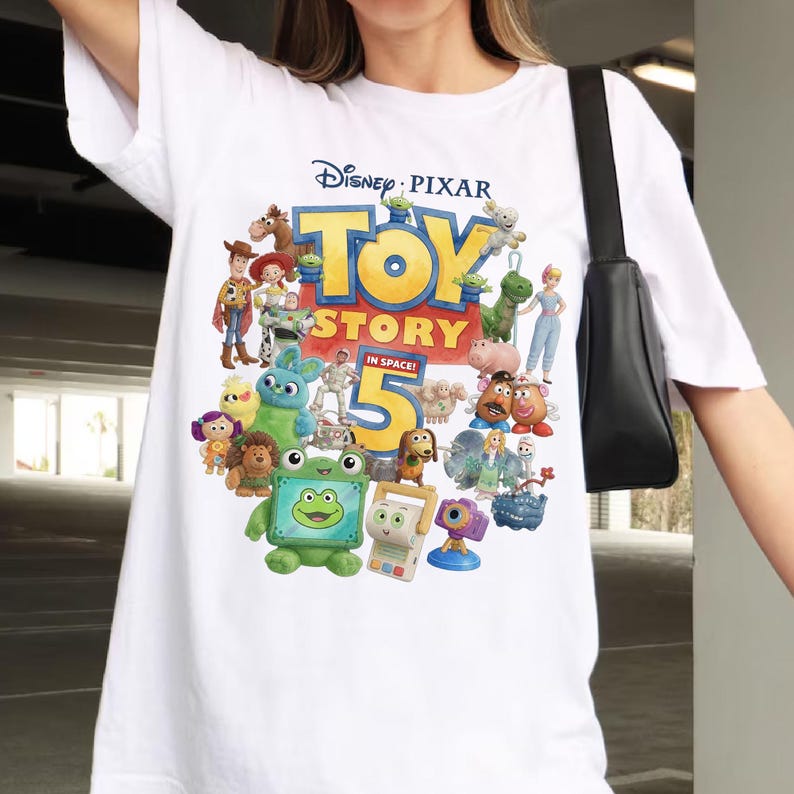 Op de afbeelding: Een wit T-shirt met korte mouwen en een kleurrijke grafische print van diverse geanimeerde personages uit de Disney Pixar-film Toy Story 5 In Space. Het ontwerp bevat de filmtitel en een groot cijfer vijf in het midden van het shirt.