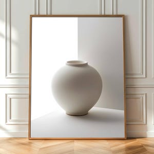May include: A framed print featuring a minimalist still life. The artwork showcases a large, round, off-white vase with a wide opening. The vase is set against a white and light gray backdrop, framed in a gold-colored border.