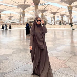 Könnte beinhalten: Eine Person trägt einen dunkelbraunen Hijab und eine Sonnenbrille und posiert in einem großen Außenbereich. Der Hijab hat die gleiche Farbe wie das lange, fließende Kleid. Der Hintergrund zeigt einen großen, offenen Bereich mit architektonischen Details.