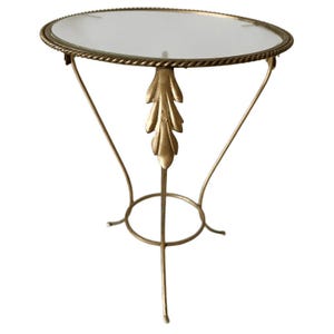 Può includere: Un tavolino in metallo color oro con piano circolare in vetro e bordo a corda. La base è composta da tre gambe sottili e curve collegate da un anello di supporto, con un elemento decorativo a forma di foglia su ciascuna gamba.