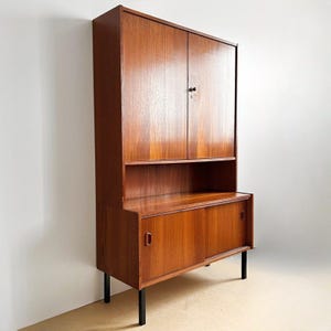 Könnte beinhalten: Ein hoher Holzschrank mit einer warmen, braunen Holzmaserung. Der obere Bereich verfügt über zwei Flügeltüren mit einem kleinen zentralen Knopf, während der untere Teil zwei Schiebetüren mit eingelassenen Griffen umfasst, getragen von vier schwarzen Beinen.