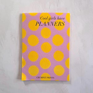 May include: A rectangular planner with a light purple cover featuring a pattern of bright yellow polka dots. The text "Cool girls have PLANNERS" is printed in black near the top, and "CRUMPET PRINTS" is printed in black at the bottom.