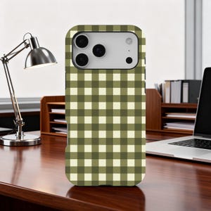 May include: A phone case with a green and cream checkered pattern. The case is on a wooden desk with a desk lamp and a laptop in the background. The phone case has a camera and flash.