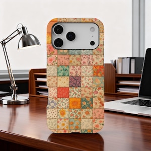 May include: A floral patchwork phone case featuring a variety of colorful floral patterns in squares. The case has a camera cutout and button placements. The case is on a wooden desk with a desk lamp and laptop in the background.