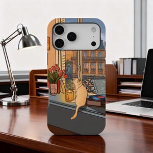 May include: A phone case depicting a cat gazing out of a window overlooking a European city. The case has a warm colour scheme, with a record player, tulips, and a lit candle on the windowsill. A desk lamp and laptop are also visible.