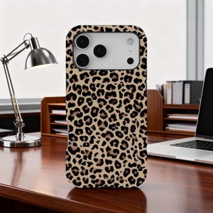 Puede incluir: Una funda de teléfono con un diseño de estampado de leopardo. La funda tiene un fondo beige con manchas marrones oscuras. La funda del teléfono está sobre un escritorio de madera con una lámpara de escritorio y un portátil en el fondo.