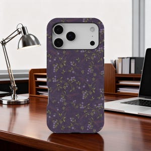 May include: A purple phone case with a floral pattern featuring small purple flowers and green leaves. The case is on a wooden desk with a desk lamp and a laptop in the background.