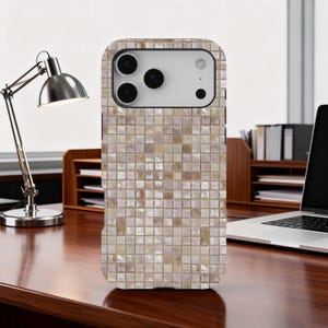 May include: A smartphone case with a mosaic tile design in shades of beige, cream, and light brown. The case has a camera cutout and button placements. The phone case is on a wooden desk with a desk lamp and a laptop in the background.