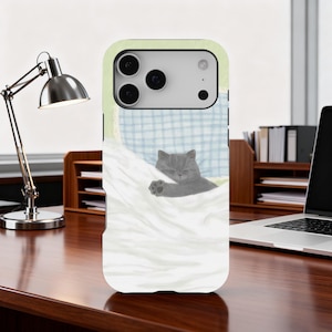 May include: A phone case featuring a gray cat sleeping under a white blanket. The cat is nestled on a blue and white checkered pillow. The case has a light green border.