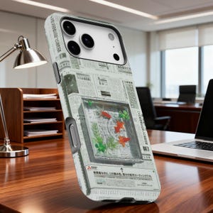 May include: A smartphone case featuring a design of a fish tank with goldfish and aquatic plants. The case has a newspaper print background with Japanese text. The case is on a wooden desk.