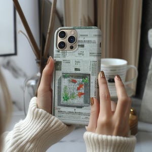 May include: A smartphone case with a unique design featuring a fish tank scene. The case has a newspaper-like background with a clear window showcasing orange fish and green plants. The case is held by a person wearing a white sweater.