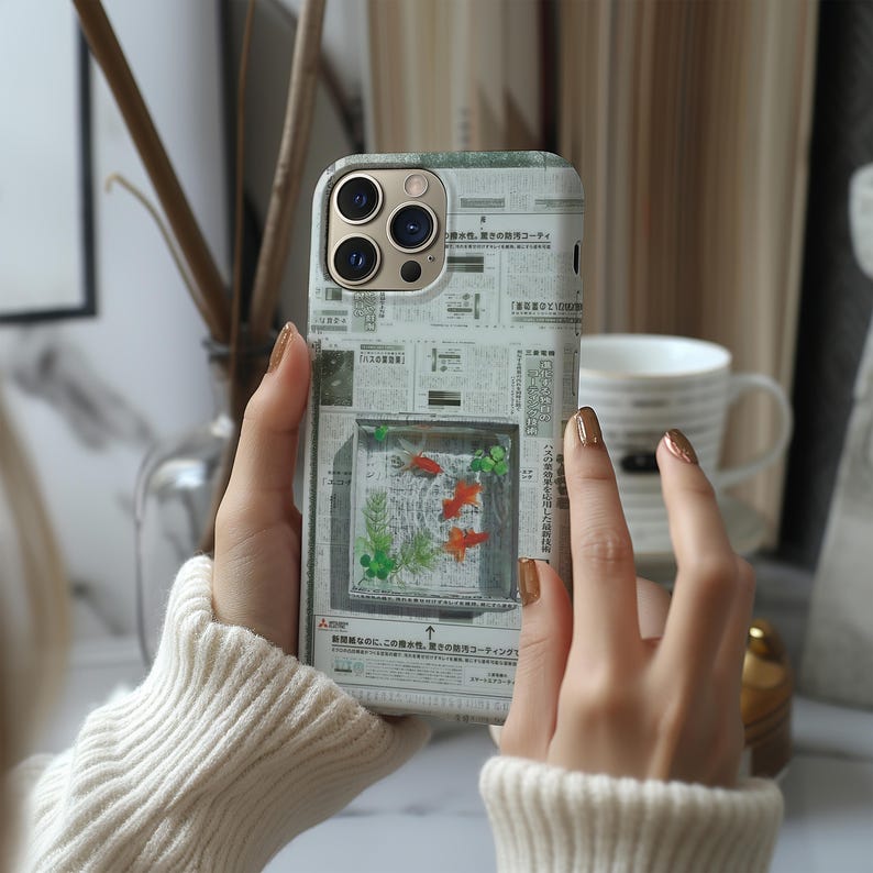 May include: A smartphone case with a unique design featuring a fish tank scene. The case has a newspaper-like background with a clear window showcasing orange fish and green plants. The case is held by a person wearing a white sweater.