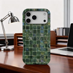Pode incluir: Uma capa de telefone com mosaico verde com um recorte para a câmera. A capa tem um padrão de azulejos quadrados em tons de verde, azul e toques de amarelo. A capa está em uma mesa de madeira.