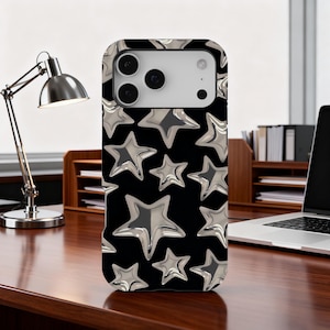 Może przedstawiać: Czarne etui na telefon z wzorem srebrnych, metalicznych gwiazd. Gwiazdy różnej wielkości są równomiernie rozmieszczone na etui. Etui leży na drewnianym biurku, w tle lampa biurkowa i laptop.