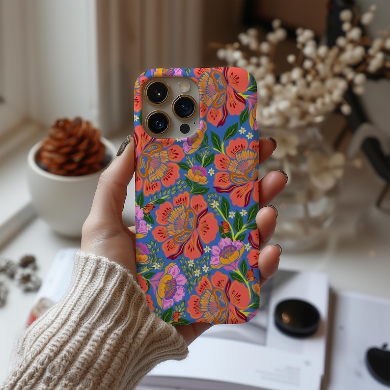 May include: A floral phone case featuring a vibrant pattern of orange, pink, and yellow flowers on a blue background. The case has a protective design with cutouts for the camera and buttons. The floral design is repeated across the entire surface of the case.