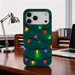 May include: A dark green phone case featuring a festive design of colorful string lights. The lights are in various colors, including red, yellow, green, and purple, with small dots scattered across the surface. The case is on a wooden desk.