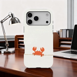 Peut inclure: Coque de téléphone blanche avec un dessin de crabe orange. Le crabe de dessin animé a des yeux noirs et est dessiné sur une fine ligne horizontale. La coque a une finition brillante.
