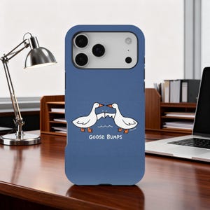 May include: A blue phone case with a cartoon design of two white geese, beak to beak, above the words "Goose Bumps". The case has openings for the phone's camera and buttons.