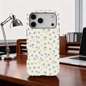 May include: A phone case featuring a floral design. The case has a cream-coloured background with a repeating pattern of small blue flowers and green leaves. The phone case is on a wooden desk.