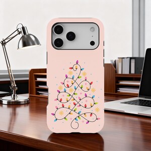 May include: A light pink phone case featuring a Christmas tree design made of colorful string lights and yellow stars. The phone case has a smooth finish and is designed to fit a smartphone.