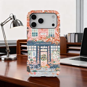 May include: A phone case with a detailed illustration of a bookstore. The design features a pink brick building, a blue awning, and a bicycle. The text "THE BOOKSHOP" is above the entrance, with "EST 2024" below. The case has a floral pattern.