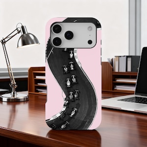 May include: A phone case featuring a black and white race track design with several race cars. The case has a pink background and is designed to fit a smartphone. The case is on a wooden desk.
