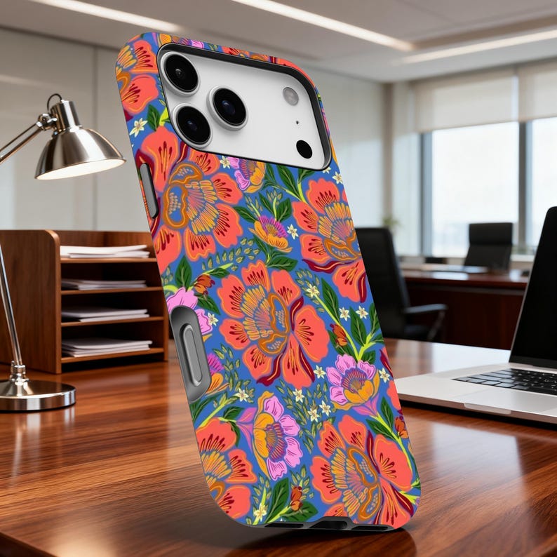 May include: A floral patterned phone case featuring a vibrant design of large orange and pink flowers on a blue background. The case has a black camera housing and a silver-colored side button. The phone case is on a wooden desk.