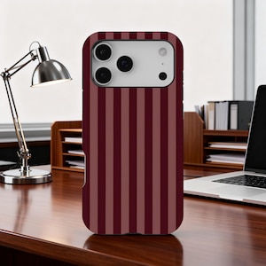 May include: A burgundy and maroon striped phone case. The case features a vertical stripe design and is made to fit a smartphone. The phone case is on a wooden desk with a desk lamp and laptop in the background.