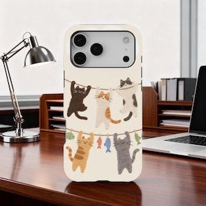 Puede incluir: Funda de teléfono con un diseño caprichoso de gatos de dibujos animados colgados de un tendedero con peces. Los gatos son de varios colores, incluyendo negro, naranja y gris, sobre un fondo crema. La funda tiene un acabado brillante.