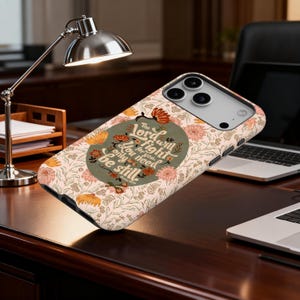 May include: A phone case with a floral pattern in peach, orange, and green. The case features the text "The Lord will fight for you. You need only be still." The case is on a dark wood surface.