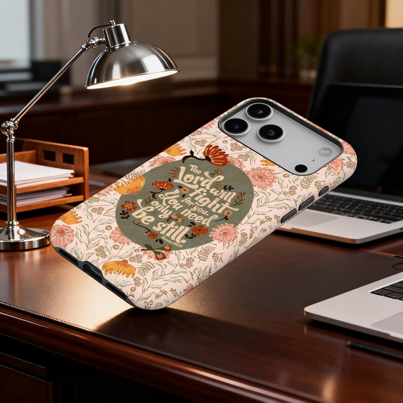 May include: A phone case with a floral pattern in peach, orange, and green. The case features the text "The Lord will fight for you. You need only be still." The case is on a dark wood surface.