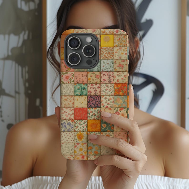 May include: A phone case with a patchwork design featuring various floral patterns in shades of pink, yellow, green, and orange. The case has a square grid pattern, with each square displaying a different floral print. The phone case is held in front of a person.