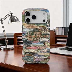 May include: A colorful phone case with a whimsical design featuring handwritten text and inspirational phrases. The case is on a wooden desk next to a laptop and a desk lamp.