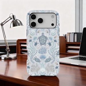 May include: A light blue phone case featuring a sea life pattern. The design includes turtles, seashells, starfish, and coral in shades of blue and gray. The case is shown on a wooden desk.