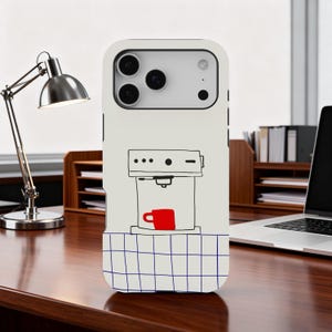 May include: A white phone case featuring a whimsical coffee machine illustration with a red coffee cup. The design includes a blue and white checkered pattern at the bottom. The case is on a wooden desk with a silver desk lamp and a laptop.