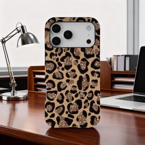 May include: A phone case with a leopard print design. The case features a tan background with black spots and gold glitter accents. The phone case is on a wooden desk.