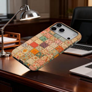 May include: A phone case with a patchwork quilt design. The case features a pattern of small, colorful squares with floral and geometric prints in shades of orange, pink, green, and cream. The case is shown on a dark wood desk.